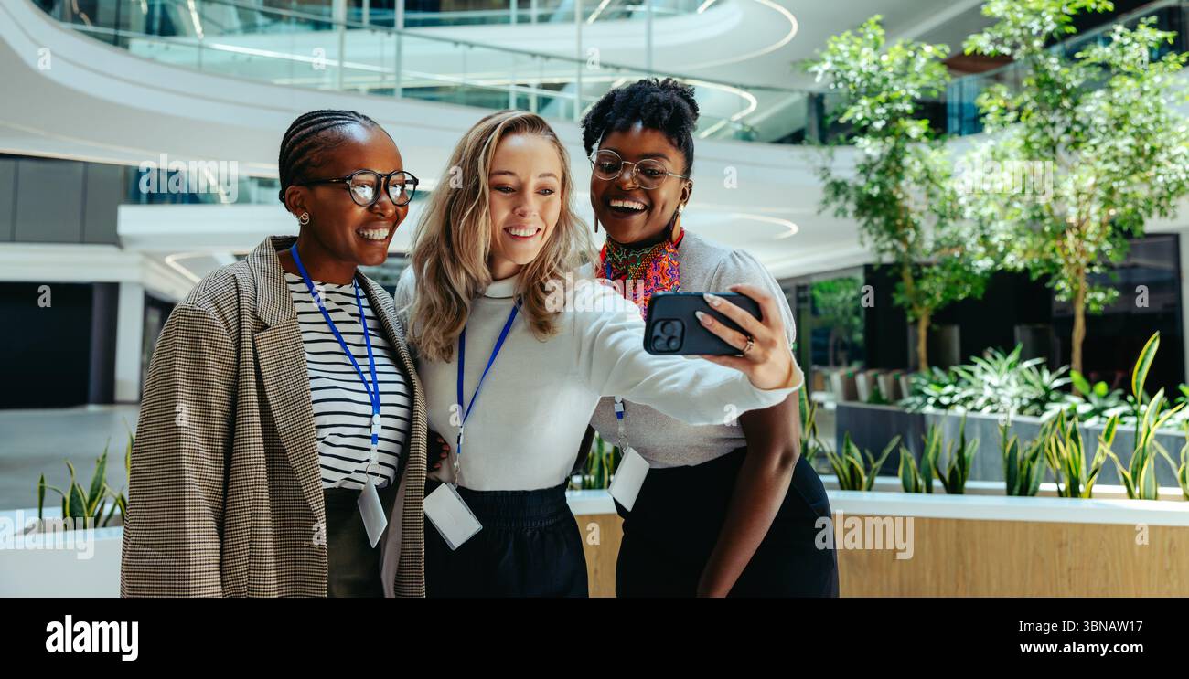Tre allegre donne d'affari scattano un selfie in un ambiente d'ufficio luminoso e moderno. L'immagine riflette temi di avanzamento di carriera, lavoro di squadra e c Foto Stock