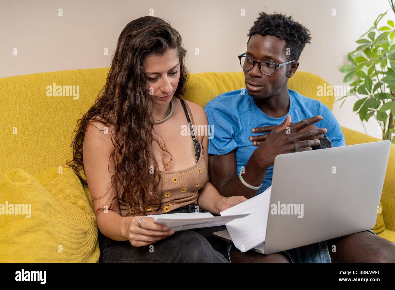 Una giovane coppia si siede su un divano e collabora alle finanze domestiche, rivedendo i documenti con un laptop. Foto Stock
