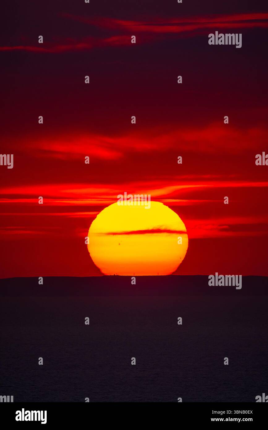 Tramonto rosso vibrante sull'orizzonte del mare con una silhouette scura di colline lontane. Foto Stock