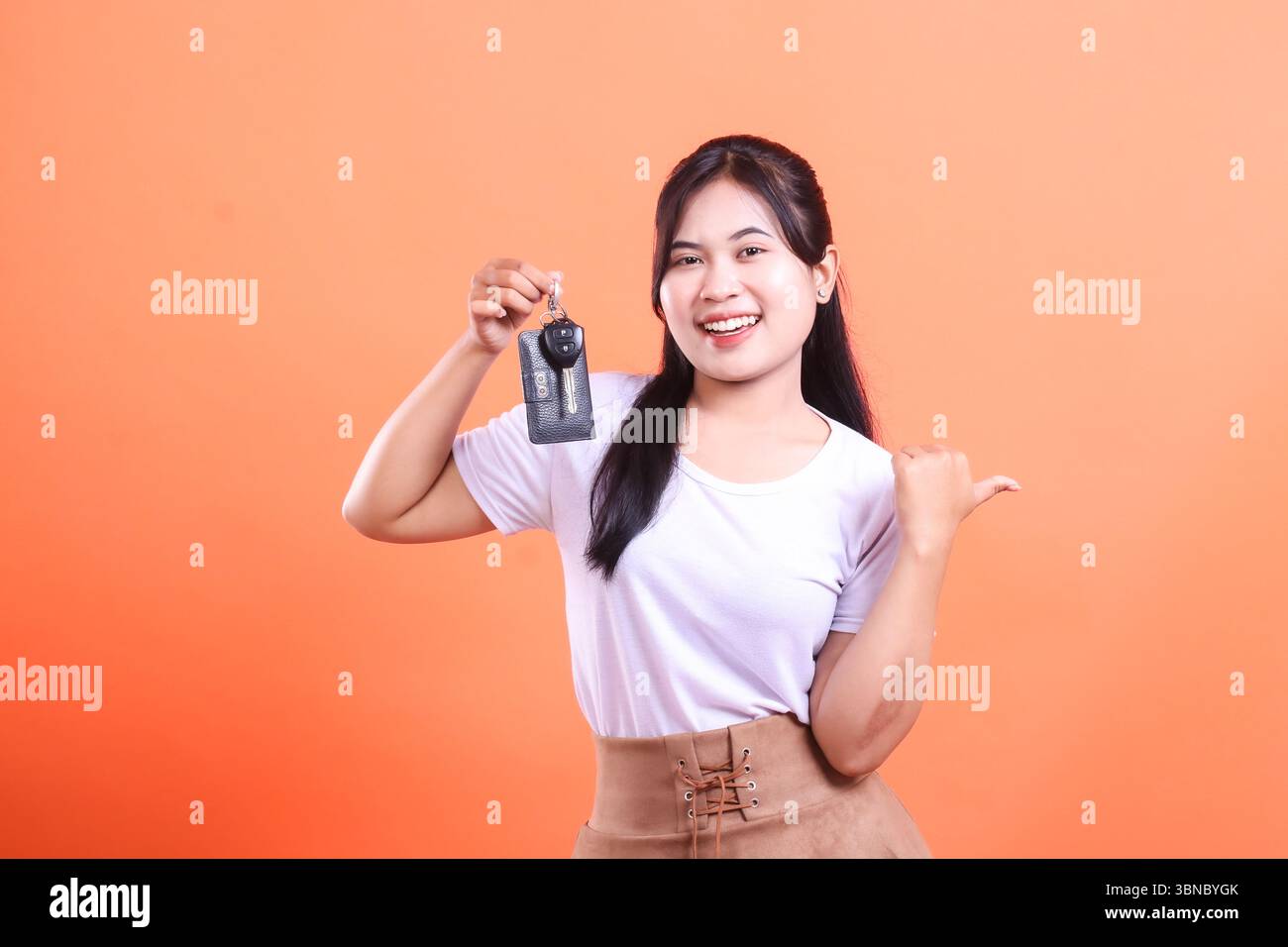Giovane donna che tiene la chiave di una macchina, sorride e mostra un pollice al suo fianco, per presentare qualcosa. isolato su sfondo arancione Foto Stock