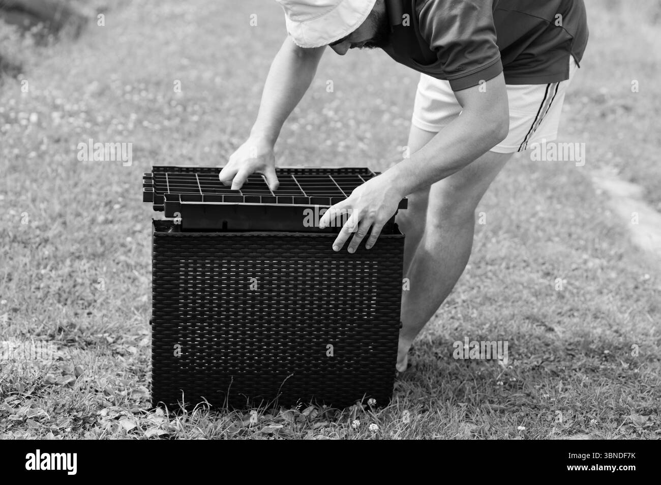 Un uomo posiziona con cura un inserto a griglia in un contenitore di stoccaggio in vimini di colore scuro. E' all'aperto sull'erba, sta assemblando il pezzo. Foto Stock
