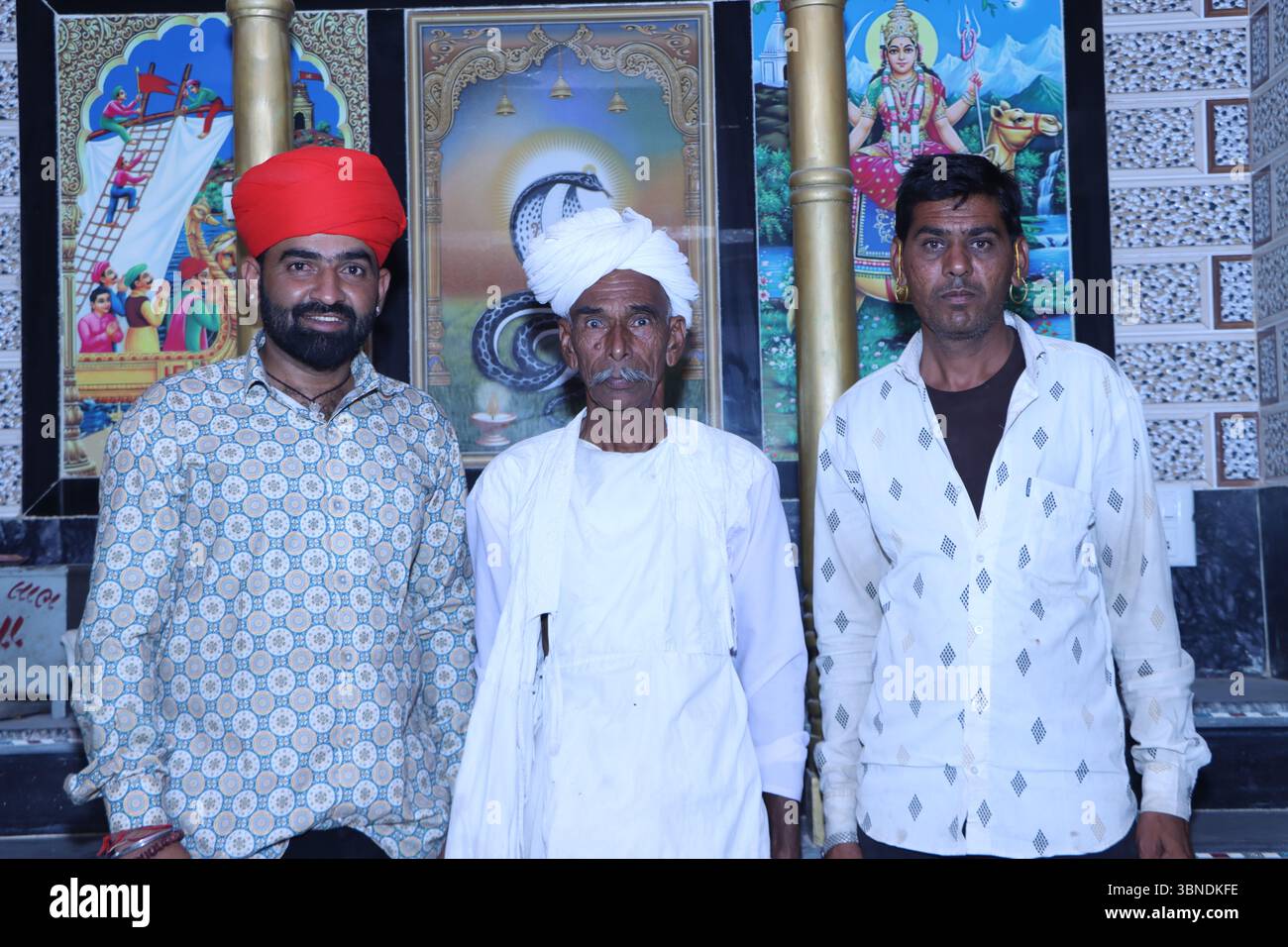 Tre uomini indiani in un abbigliamento tradizionale che posano all'interno di un tempio con poster religiosi colorati sullo sfondo Foto Stock