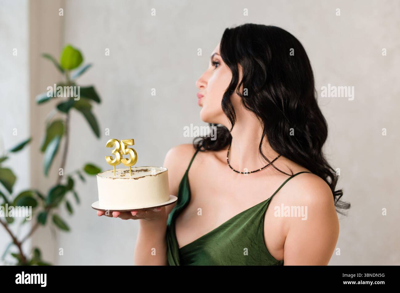 Una donna con i capelli scuri, che indossa un abito verde, tiene una piccola torta di compleanno con le candele '35'. Sta per far esplodere le candele, un momento di gioia. Foto Stock