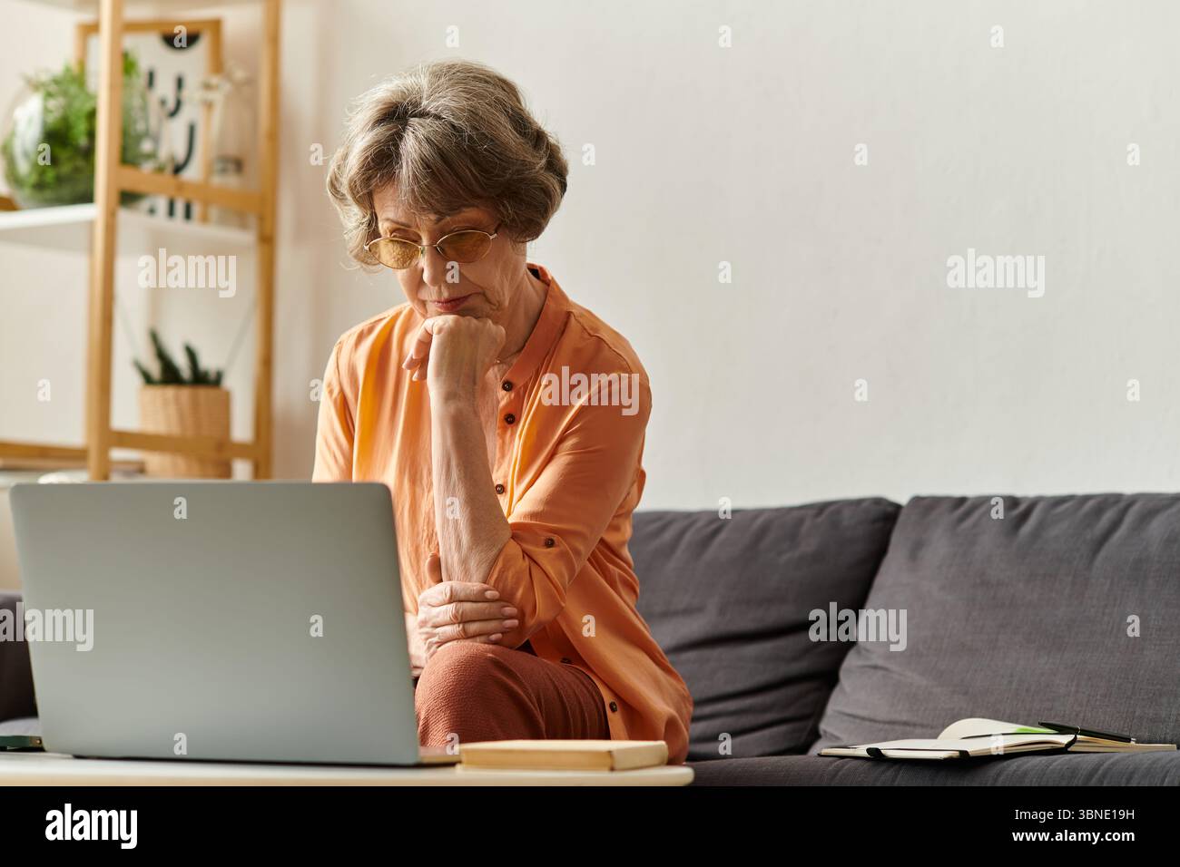 Una donna matura con un abbigliamento caldo si siede comodamente su un divano, profondamente coinvolta nello schermo del suo notebook. Foto Stock