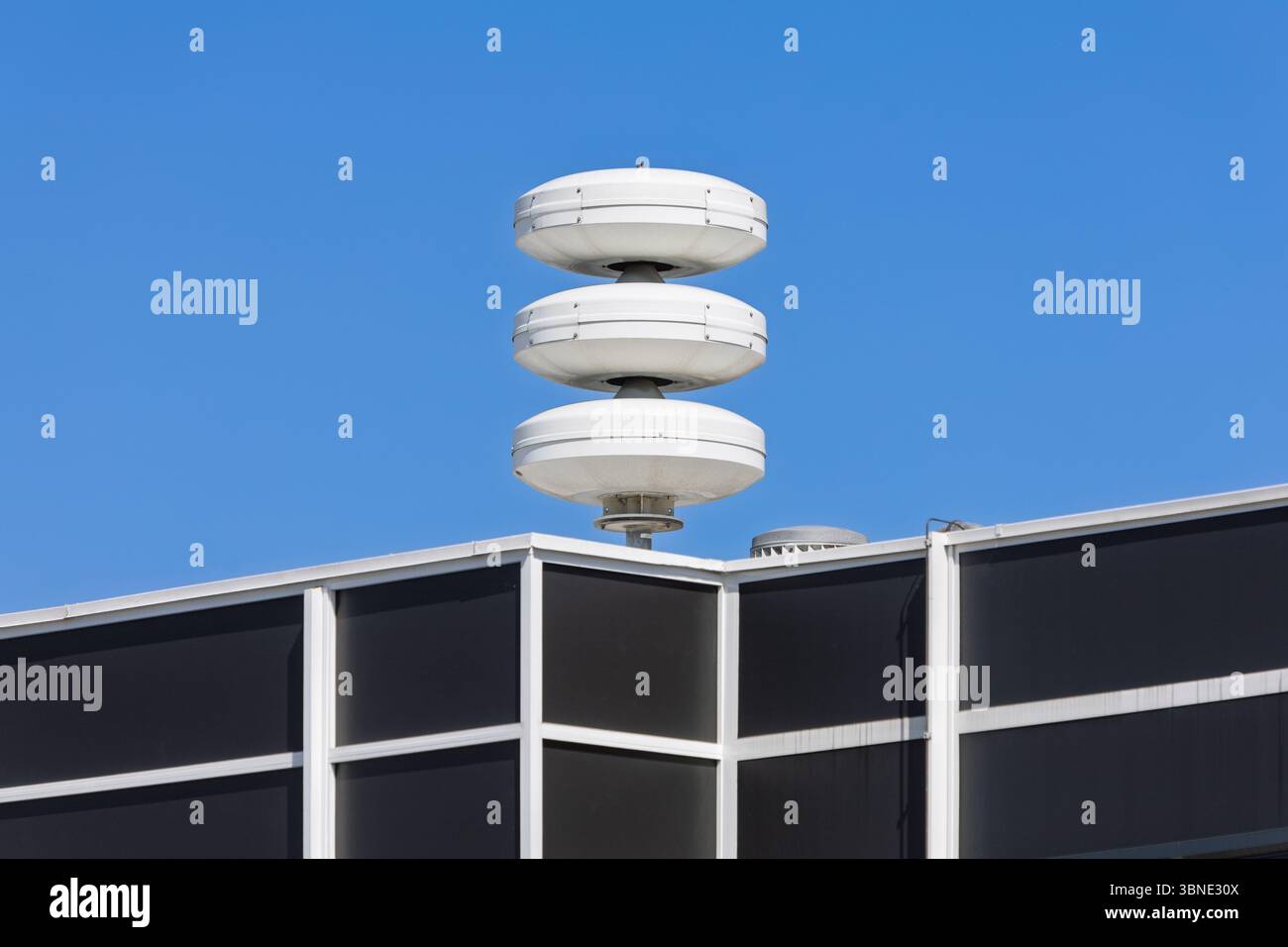 Una sirena o un sistema di allarme aereo a più livelli si erge in cima a un edificio moderno, contro un cielo azzurro, segnalando le emergenze o le comunicazioni urbane. Foto Stock
