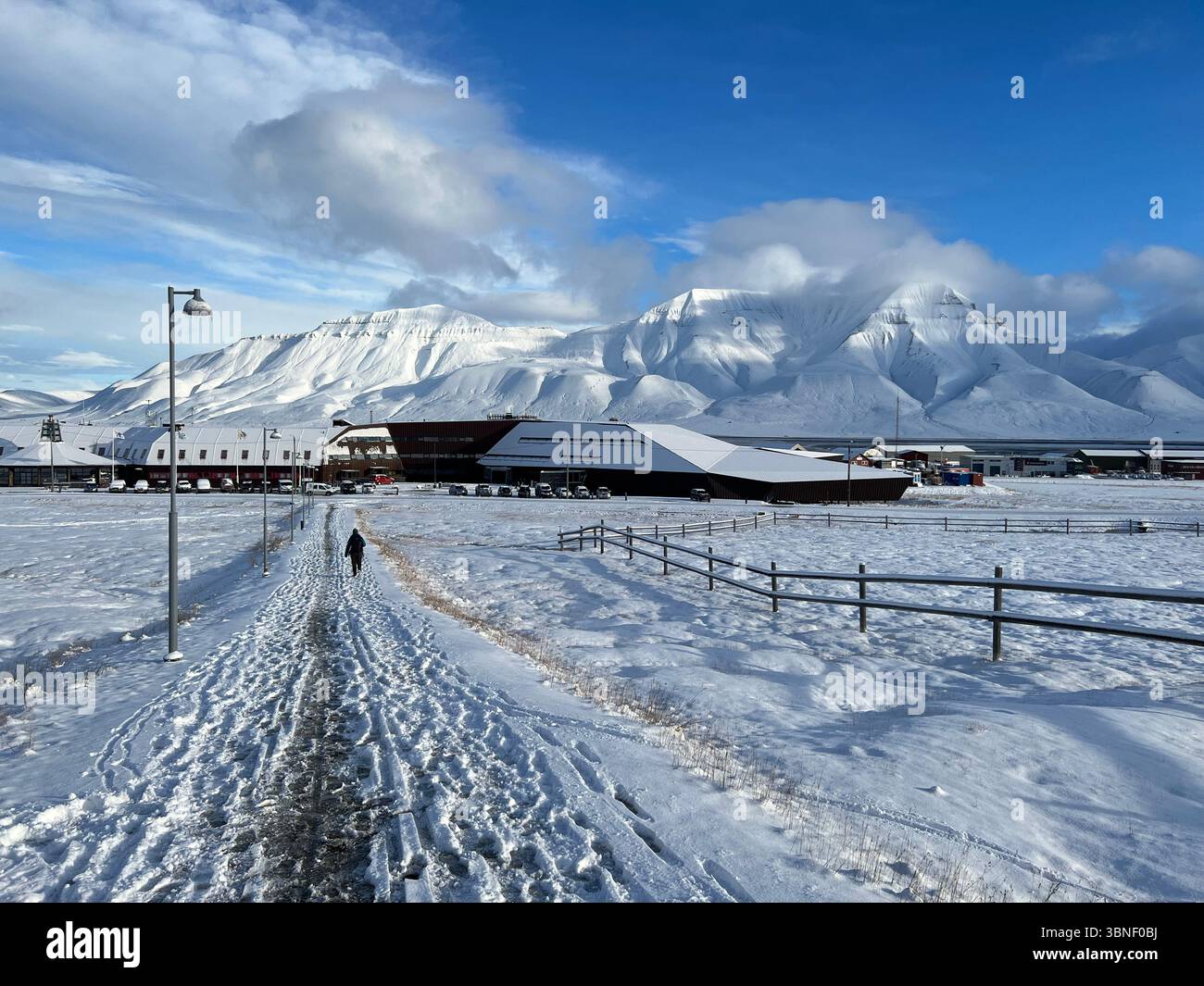 Il paesaggio invernale mostra un sentiero che conduce a montagne innevate ed edifici sotto un cielo blu brillante a Longyearbyen, Svalbard - Immagine stock catturata con smartphone
