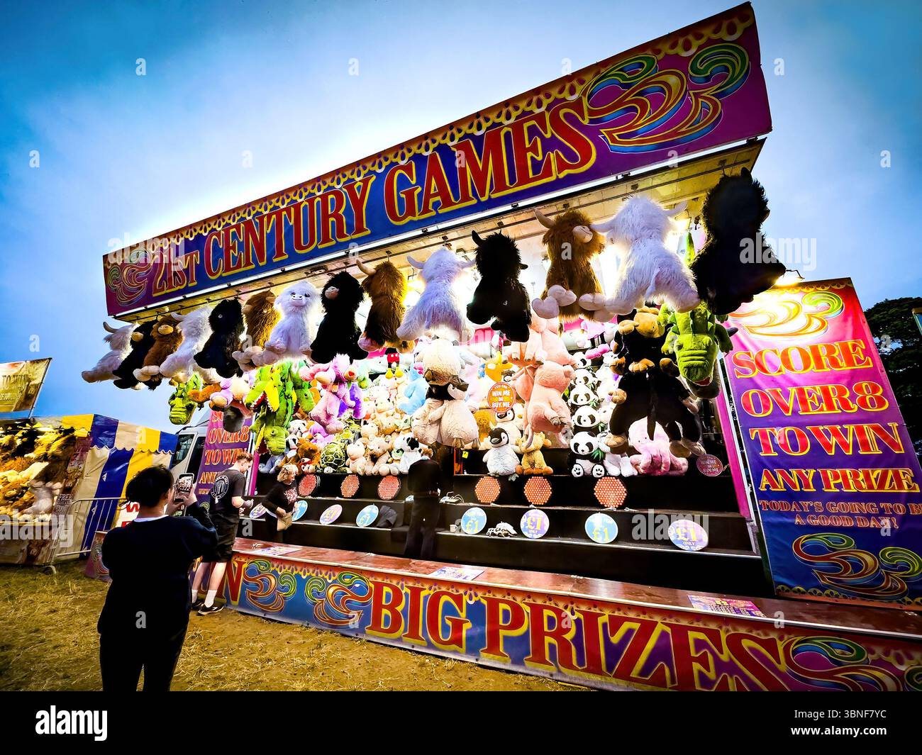 Le persone si sono riunite in uno stand luminoso al tramonto, che mette in mostra grandi animali farciti come premi per i vincitori del gioco, la città di Hoppings Moor newcastle - Immagine stock catturata con smartphone