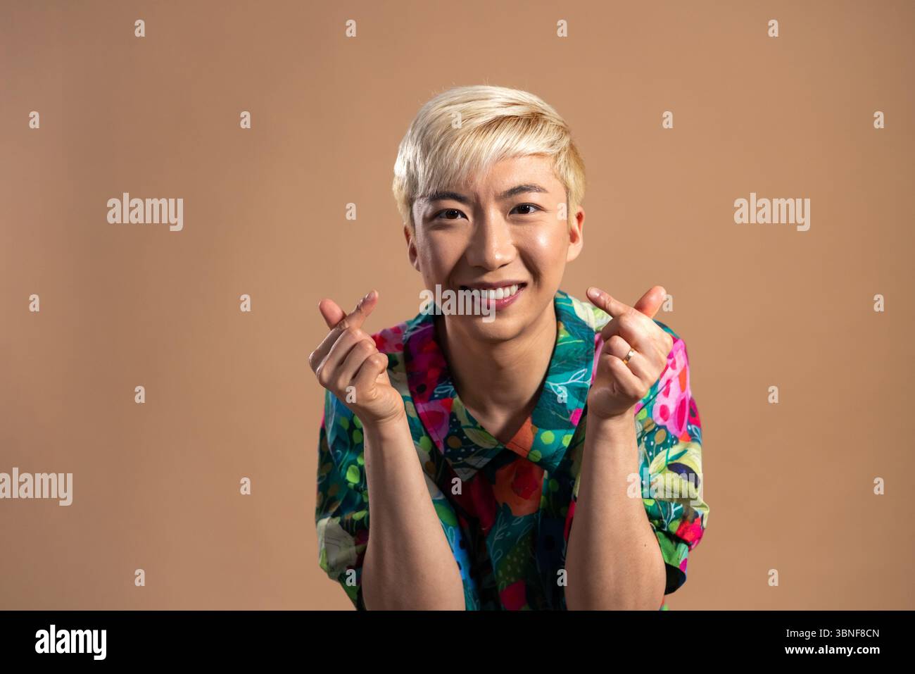 Foto in studio di un giovane maschio adulto sorridente alla macchina fotografica, che crea forme di cuore con le mani. Ha i capelli sbiancati e indossa una camicia colorata, st Foto Stock