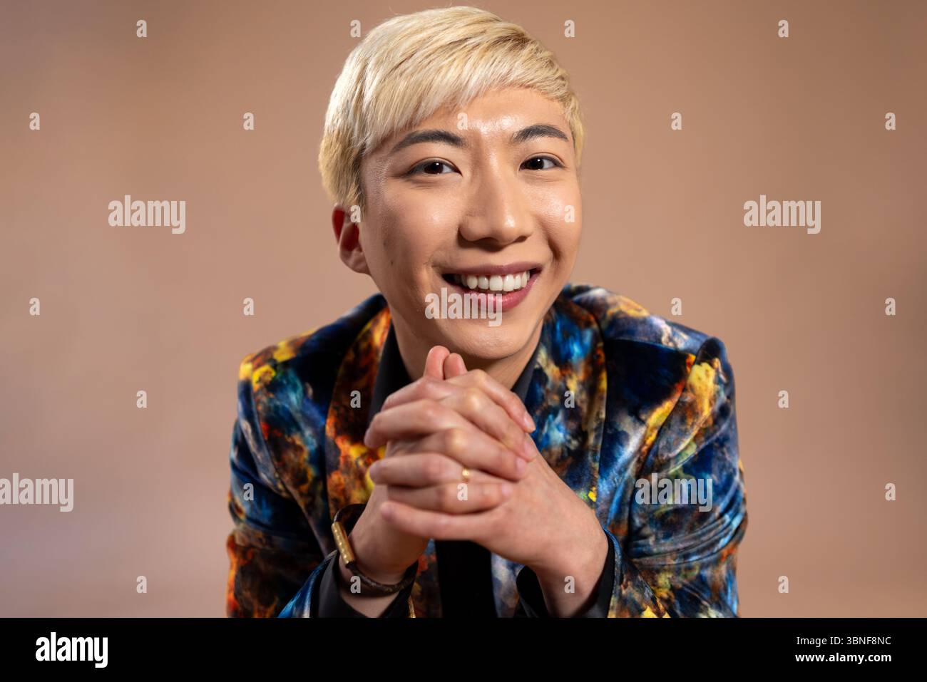 Foto in studio di un giovane maschio adulto sorridente alla macchina fotografica con le mani agganciate. Ha i capelli sbiancati e indossa un vestito di velluto colorato con la sunglasse Foto Stock