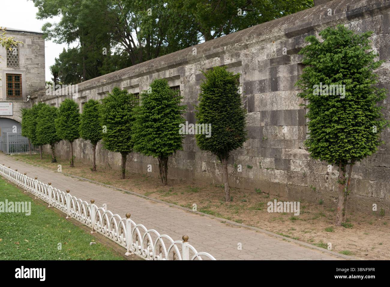 Gli arbusti verdi di forma ordinata sono allineati lungo un vecchio muro di pietra in un tranquillo parco di Istanbul, creando un'atmosfera tranquilla con luce solare intensa. Foto Stock