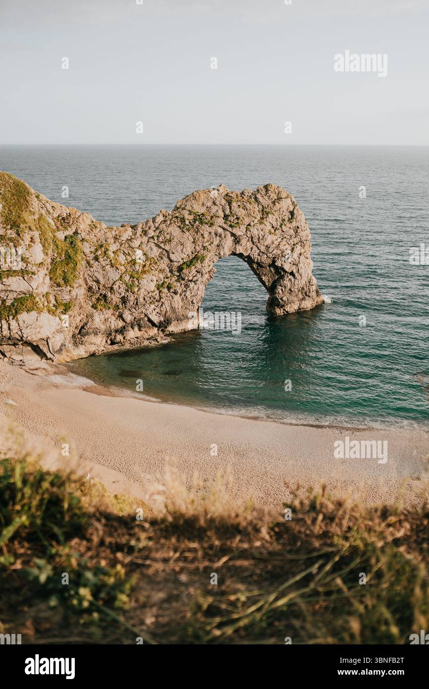 Una spiaggia rocciosa con un grande arco in acqua. L'arco è circondato da erba e l'acqua è calma Foto Stock