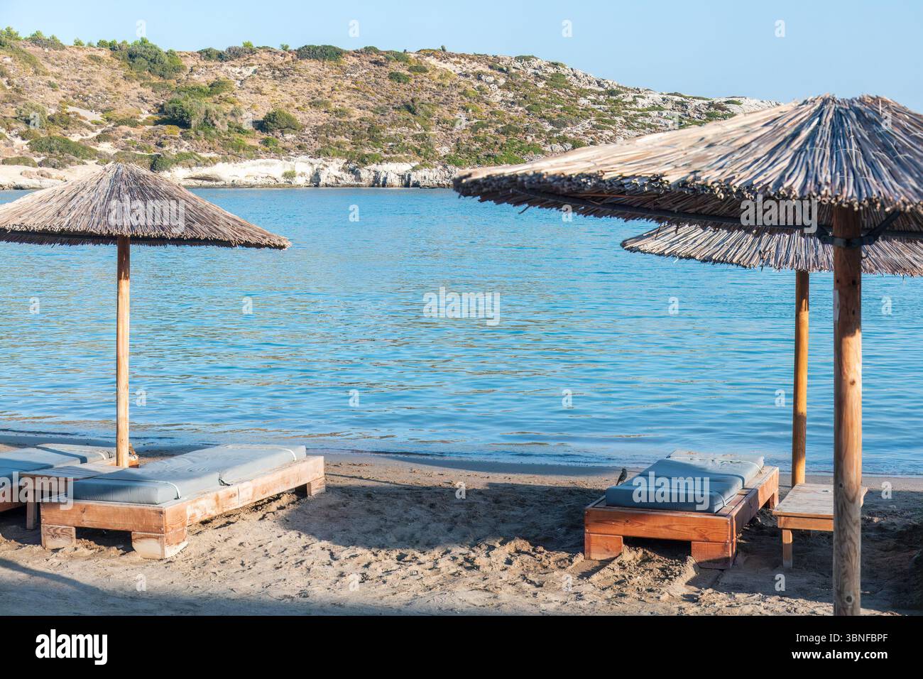Rilassante spiaggia con ombrelloni di paglia e sdraio sulla spiaggia sabbiosa. Acque turchesi tranquille e onde dolci creano un'atmosfera serena Foto Stock