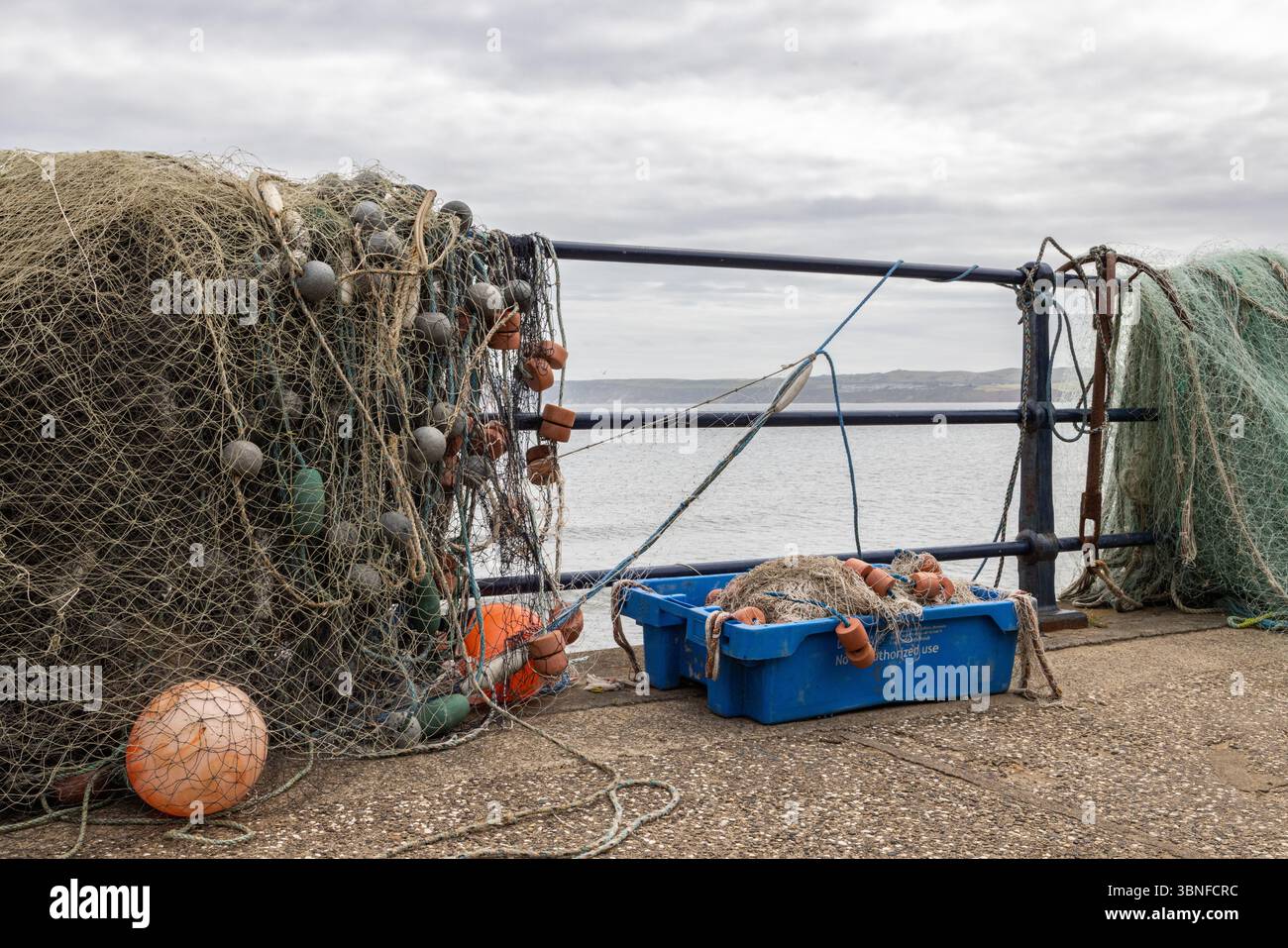 Reti da pesca, corde e carri galleggianti impilati lungo un muro del porto, simboli di una linea costiera funzionante e il volto mutevole dell'industria della pesca. Foto Stock