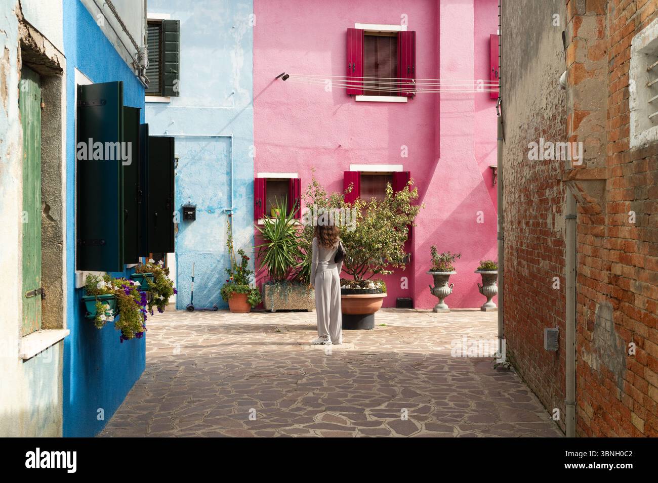 Donna in piedi in un vivace e colorato vicolo di Burano, in Italia, circondato da case dipinte con colori vivaci. Ideale per viaggi e destinazioni uniche. Foto Stock