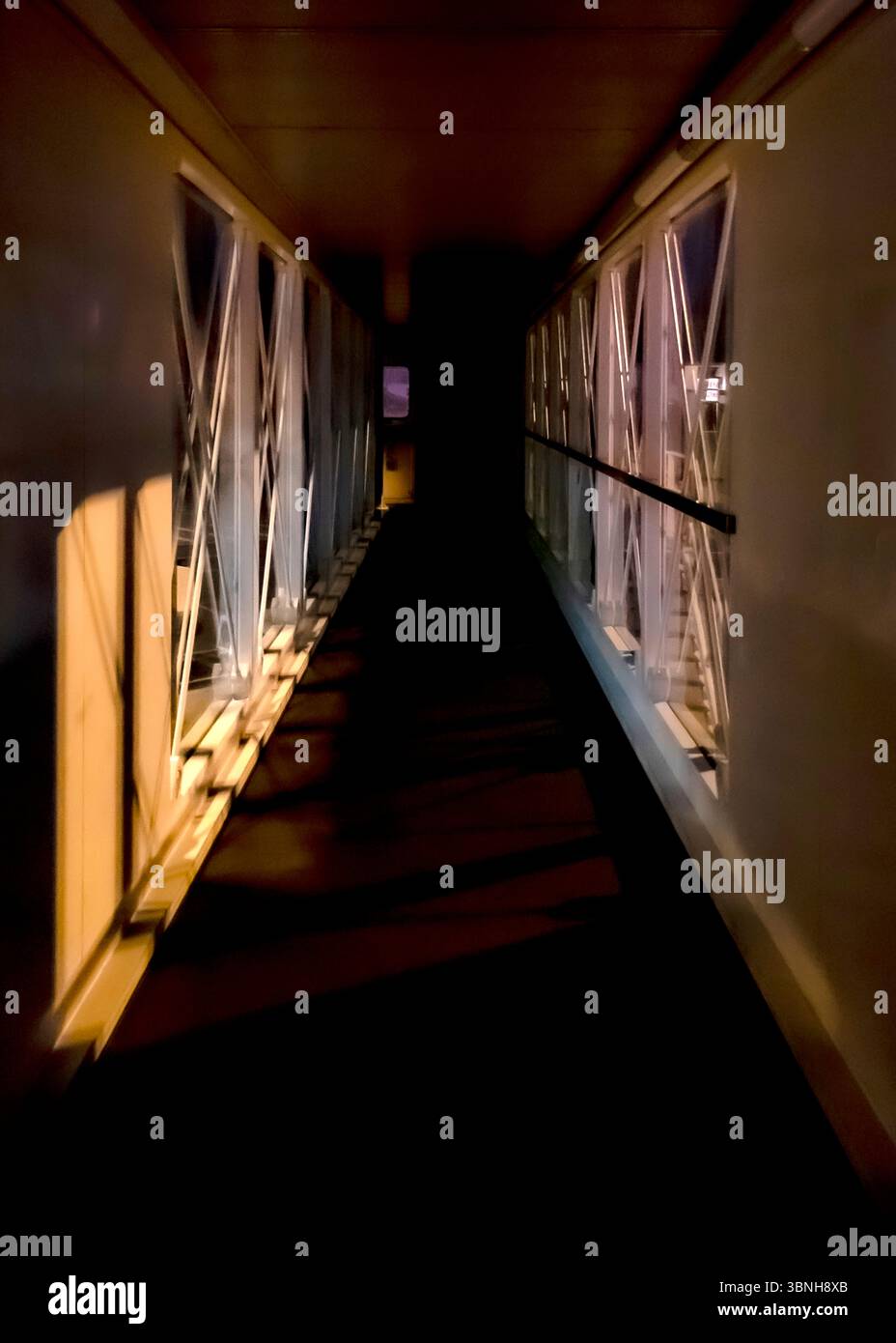 Un corridoio lungo e scuro con finestre su entrambi i lati. Il corridoio è poco illuminato, con ombre proiettate dalle finestre. La fine del corridoio è oscurata da Foto Stock