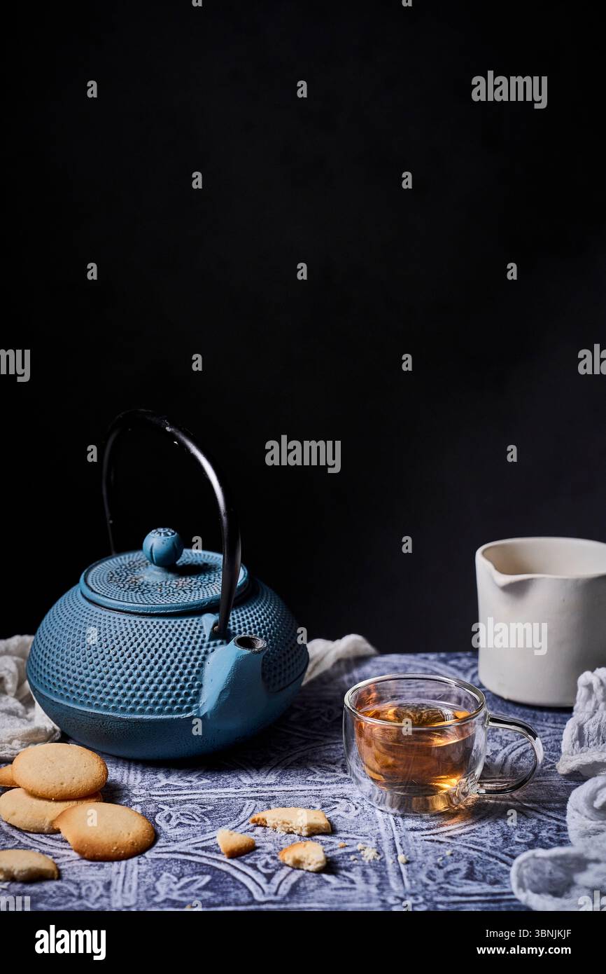 Un'accogliente preparazione per il tè con una teiera blu, una tazza di tè fumante e deliziosi biscotti su un panno a motivi geometrici. Una caraffa per il latte aggiunge un tocco di eleganza Foto Stock