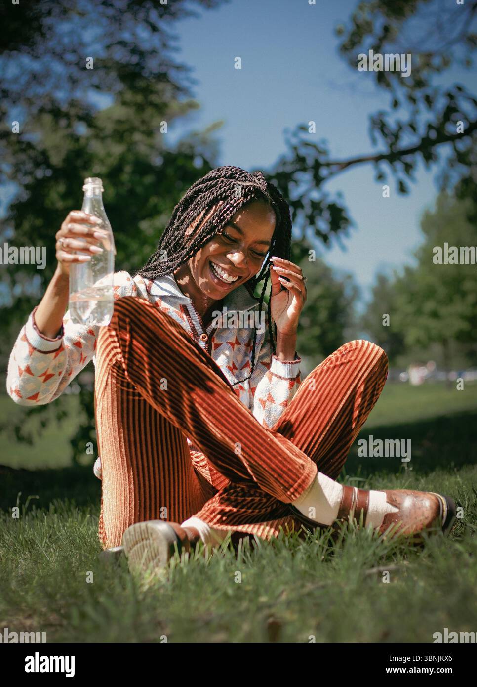 Una gioiosa donna afroamericana con i capelli intrecciati ride di cuore mentre si siede sull'erba e tiene in mano una bottiglia d'acqua. Il sole splende brillantemente, aggiungendo W. Foto Stock