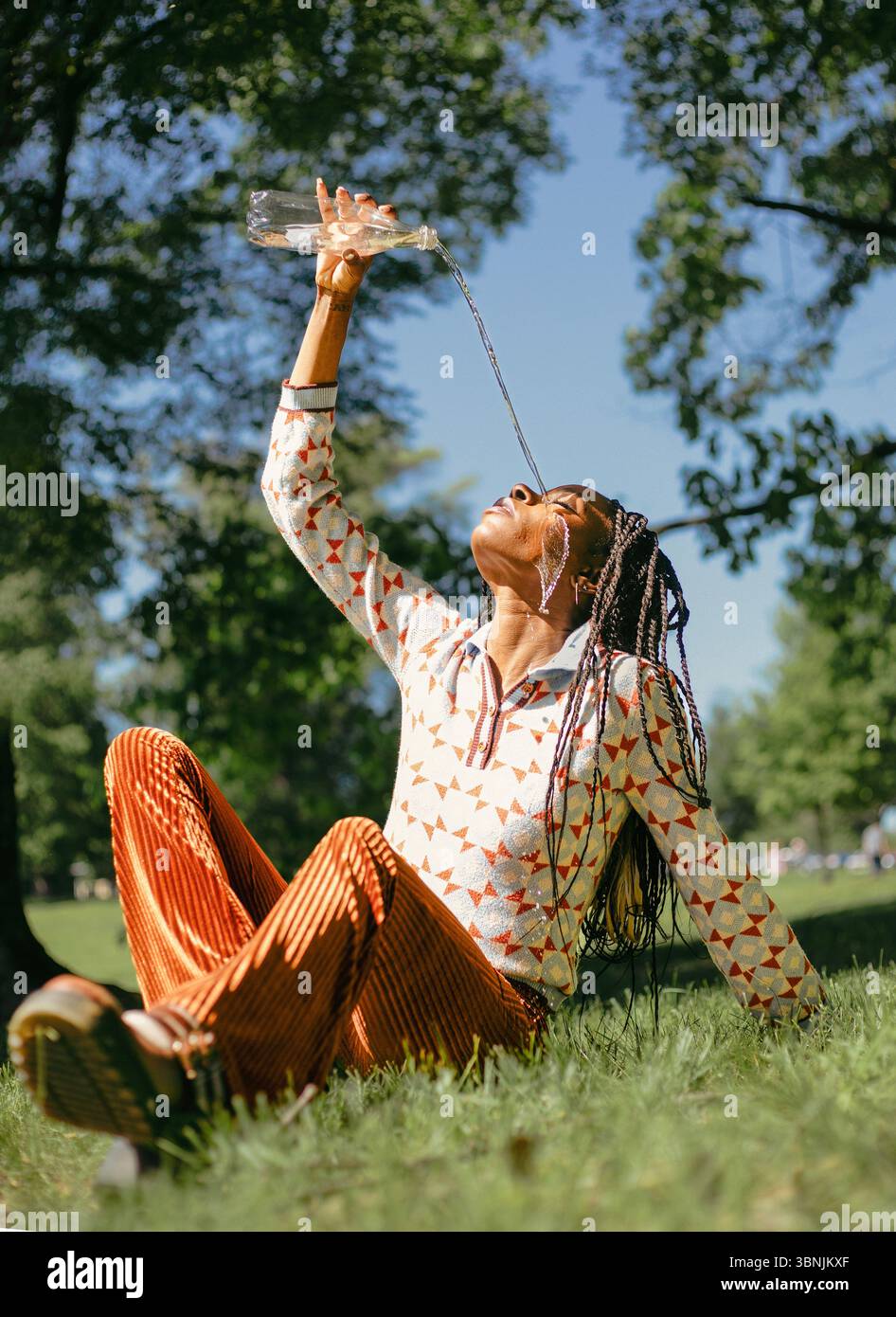 Una donna afroamericana con i capelli intrecciati ama l'acqua in natura, seduto sull'erba in un parco illuminato dal sole. Versa l'acqua da una bottiglia con grazia, apprecia Foto Stock