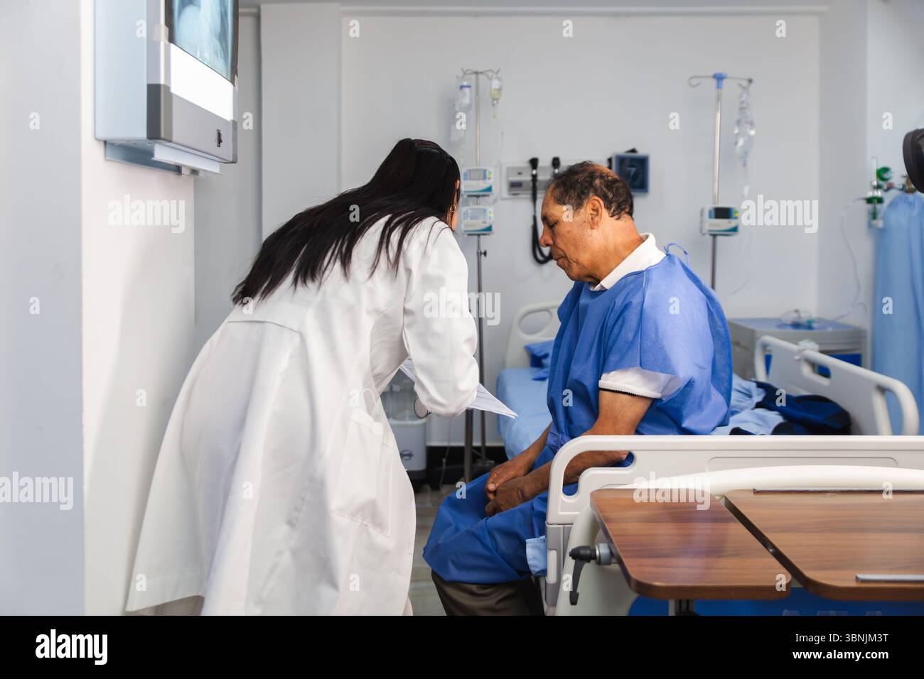 Un medico donna in un cappotto bianco si consulta con un paziente anziano di sesso maschile seduto su un letto d'ospedale, discutendo informazioni mediche in un luminoso e ben attrezzato Foto Stock