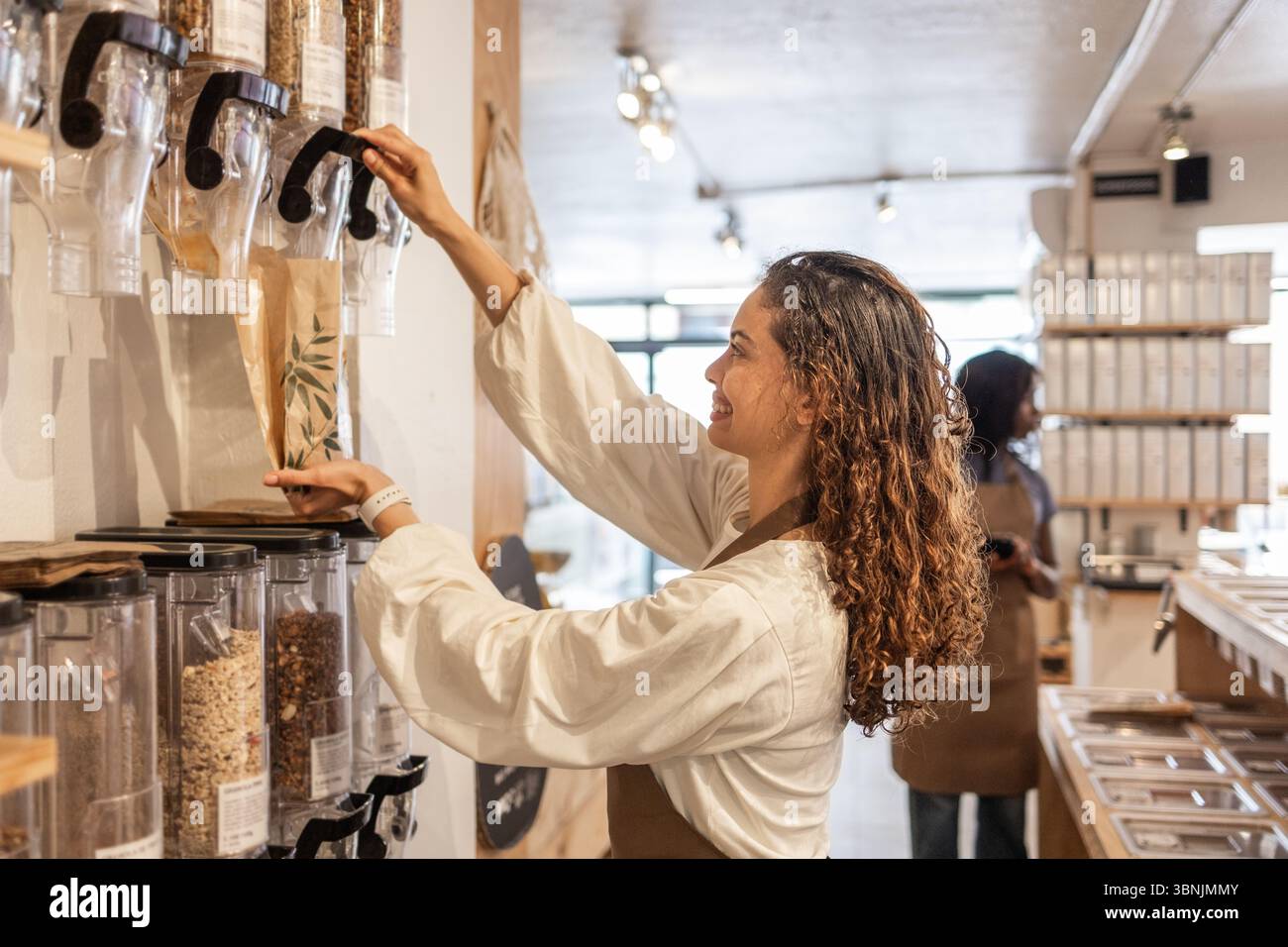 Un impiegato sorridente riempie un sacchetto di carta con grani provenienti dai dispenser nella sezione bulk di un supermercato biologico. Gli scaffali sullo sfondo suggeriscono un pozzo Foto Stock