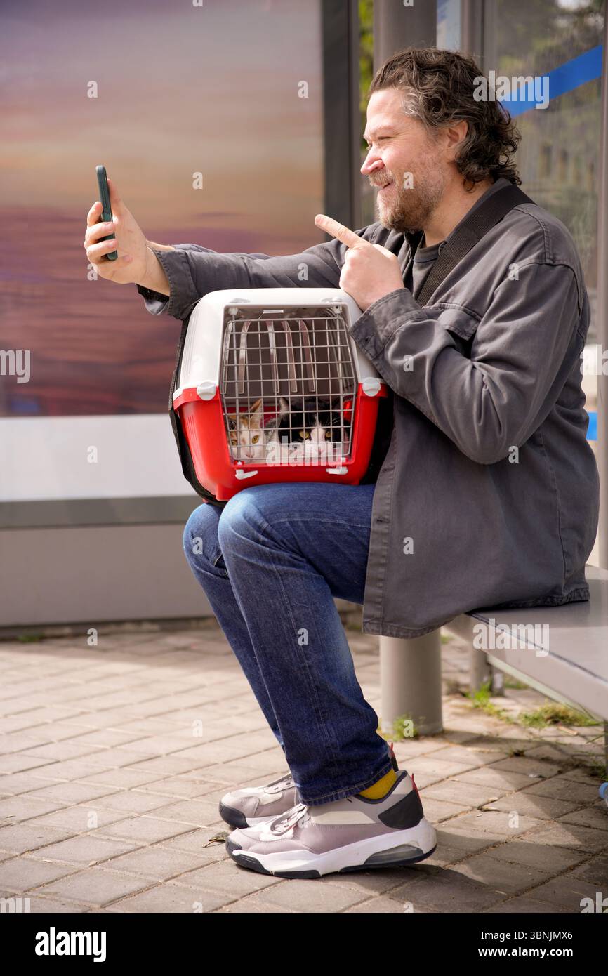 Un uomo si siede alla fermata dell'autobus con due gatti in braccio, scattando un selfie con il suo smartphone. La scena cattura un momento leggero in urba Foto Stock