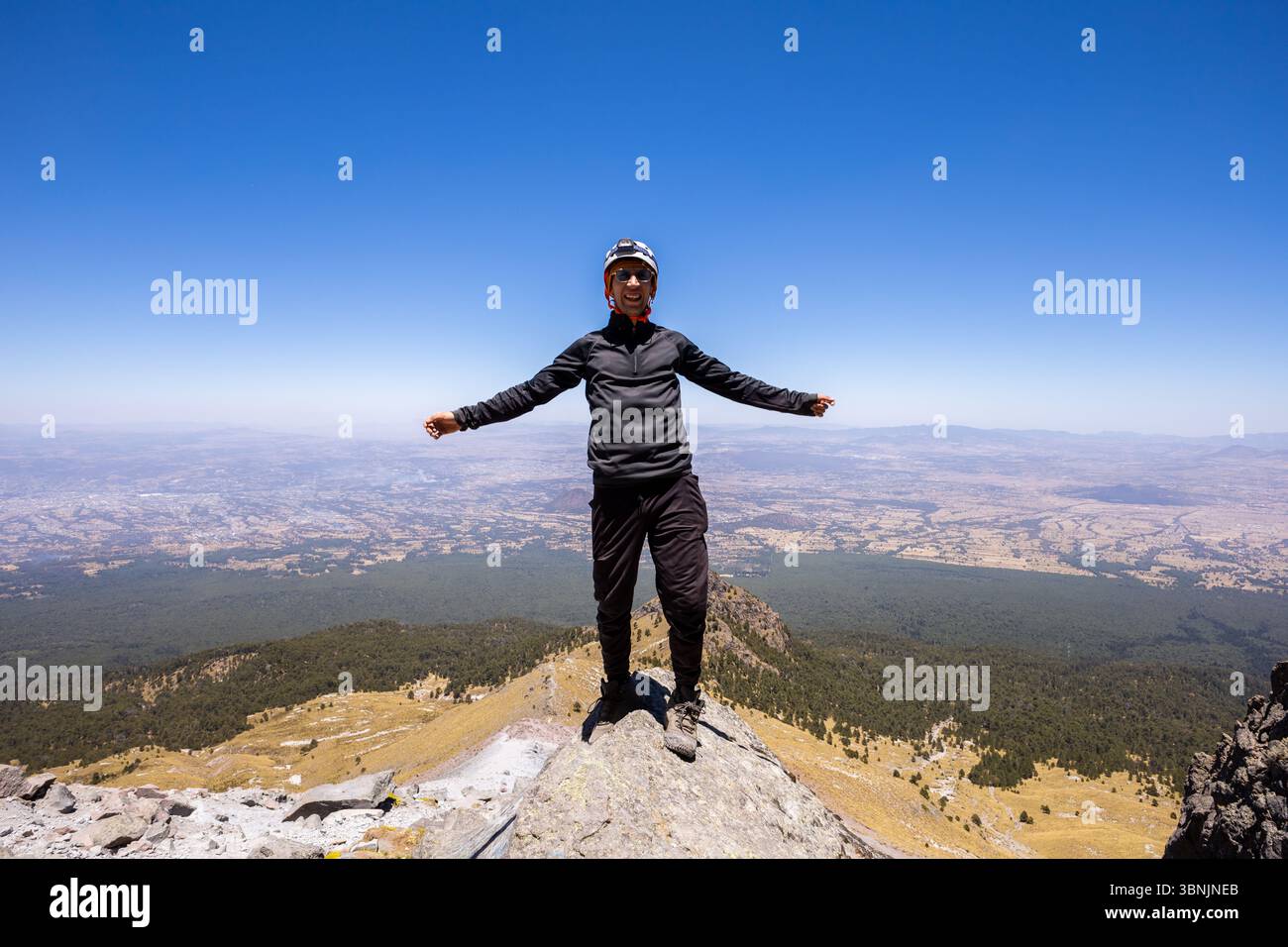 Un escursionista si erge trionfalmente in cima a una montagna, le braccia si spalancano sotto un cielo blu limpido. La vasta natura selvaggia si estende sotto, catturando l'essenza Foto Stock