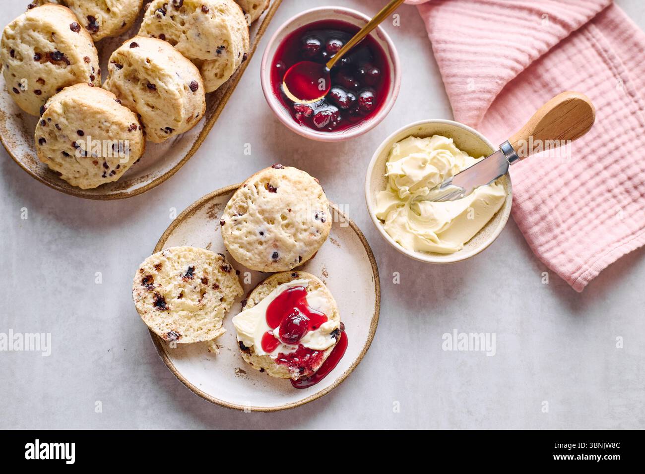 Vista dall'alto di deliziosi scones con gocce di cioccolato, ricoperti di crema coagulata e vivace marmellata di frutta, sistemati su un tavolo rustico con un tovagliolo rosa Foto Stock
