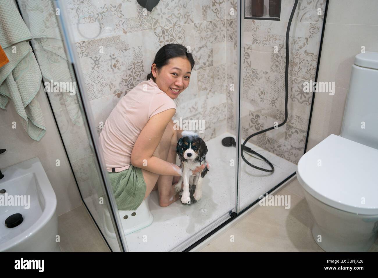 Una donna asiatica bagna con cura il suo Cavalier Re Carlo Spaniel in un bagno moderno. L'ambiente domestico è accogliente e personalizzato per il massimo comfort Foto Stock