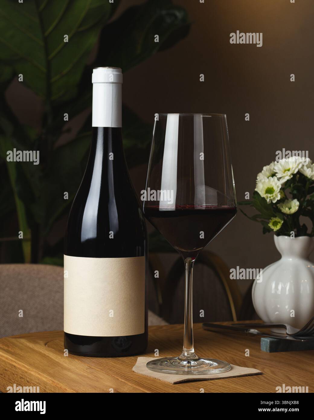 Una bottiglia di vino e un bicchiere si siedono elegantemente su un tavolo di legno in un ristorante. L'ambiente è esaltato da un vaso di fiori bianco, che crea un sofisticato dini Foto Stock