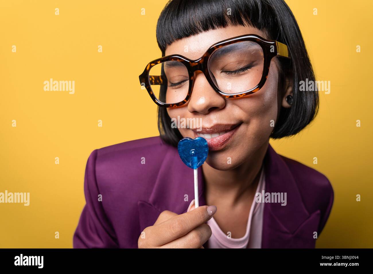 Giovane donna ispanica in giacca viola, indossa occhiali grandi, e apprezza con piacere un lecca lecca blu a forma di cuore su uno sfondo giallo vibrante, occhi Foto Stock