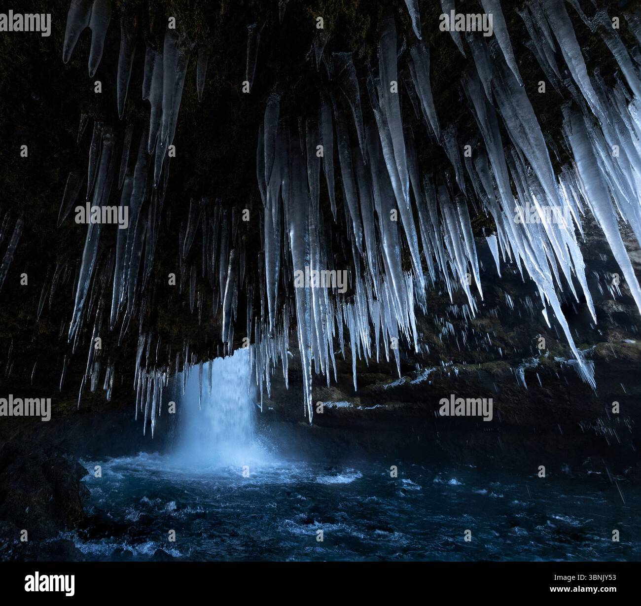 Questa immagine mistica cattura lunghi ghiacciai appesi sopra una vigorosa cascata all'interno di una grotta scura, evocando la bellezza agghiacciante del paesaggio islandese Foto Stock