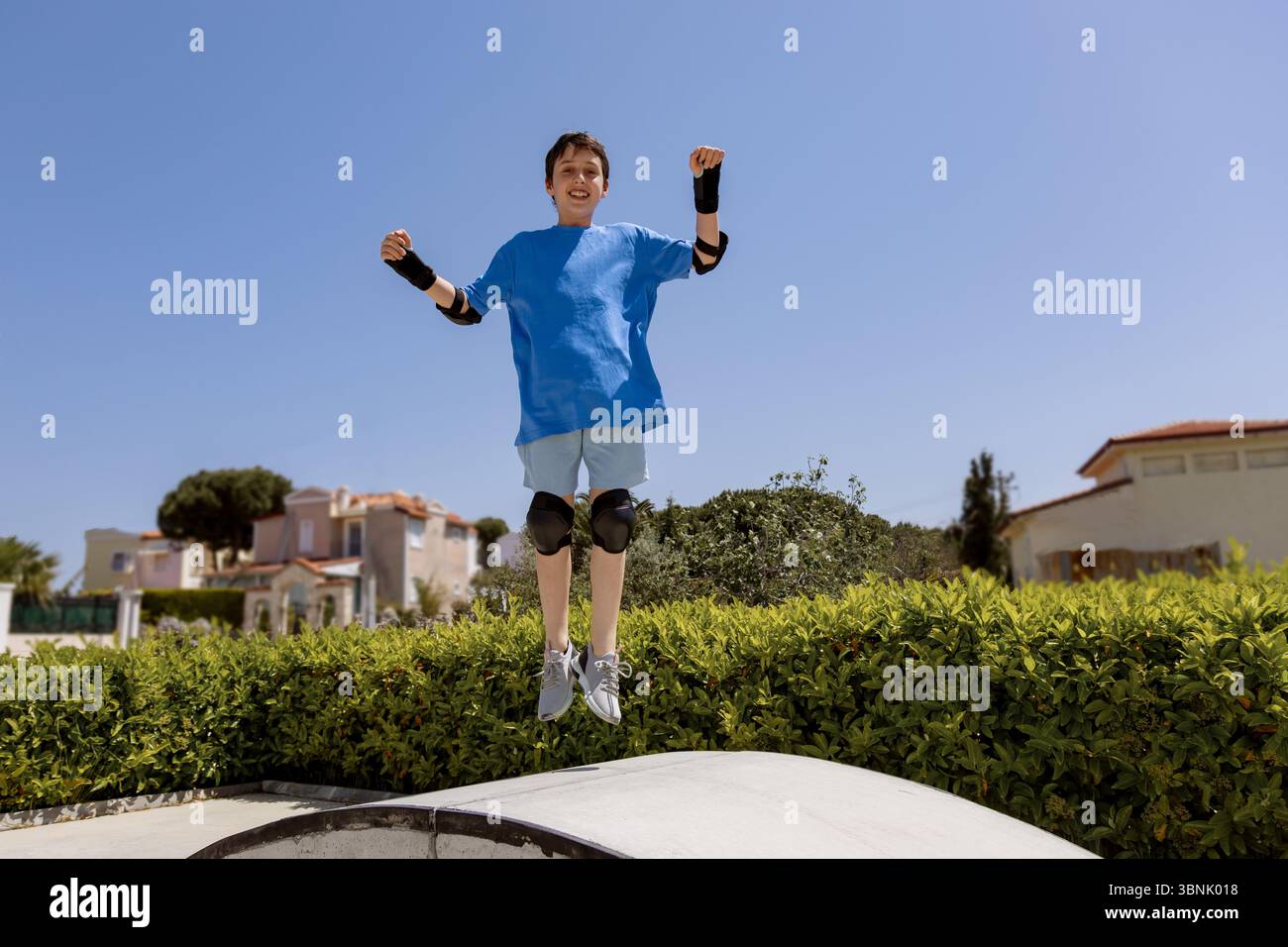 Un ragazzo gioioso che indossa una t-shirt blu e un abbigliamento protettivo salta all'aperto sotto un cielo azzurro. Le ginocchia e i gomiti garantiscono la sicurezza durante la navigazione Foto Stock