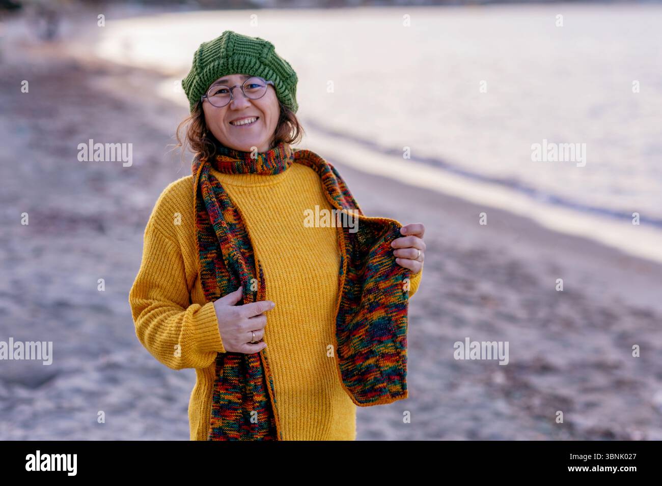 Una allegra donna di mezza età con un maglione giallo e un berretto colorato gode di un momento di pace in riva al mare al tramonto, abbracciando le casual vite costiere Foto Stock