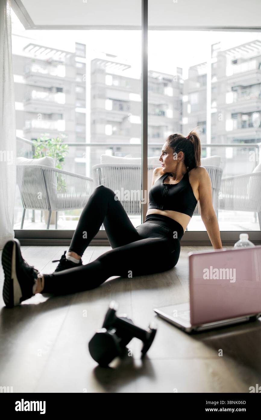 Una donna in abbigliamento sportivo nero si prende una pausa dal suo allenamento domestico, seduta sul pavimento del suo appartamento. Nelle vicinanze, un laptop e un manubrio suggeriscono il suo exe Foto Stock