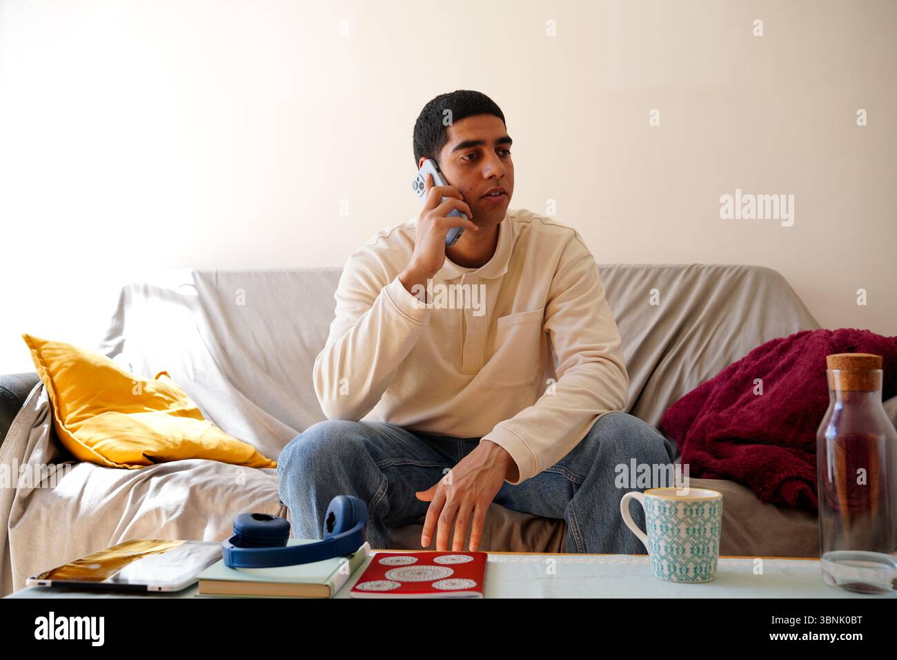 Un giovane arabo che parla al telefono, seduto su un comodo divano in casa, circondato da oggetti di uso quotidiano come libri, cuffie e una tazza di Foto Stock