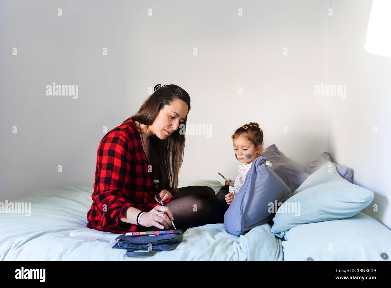 Una madre e una figlia si siedono insieme su un comodo letto a casa, impegnandosi in un creativo face painting. La calda atmosfera enfatizza il legame e l'immaginazione Foto Stock
