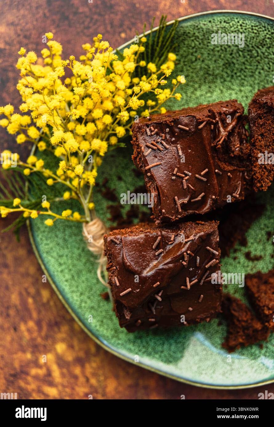 Una deliziosa torta al cioccolato ricoperta di spolverine si trova su un piatto verde, splendidamente completato da vivaci fiori gialli, adagiato su un ricco piatto marrone Foto Stock