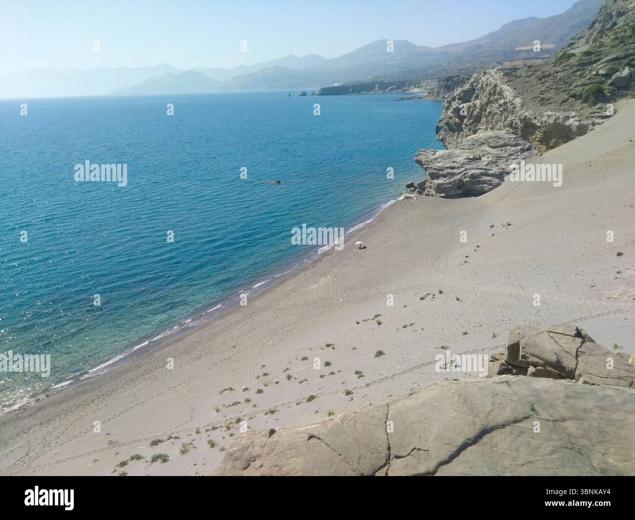 Tranquilla spiaggia sabbiosa sulla costa meridionale di Creta con acque blu e scogliere rocciose. Una singola persona sotto un ombrello aggiunge un tocco solitario alla scena. Foto Stock