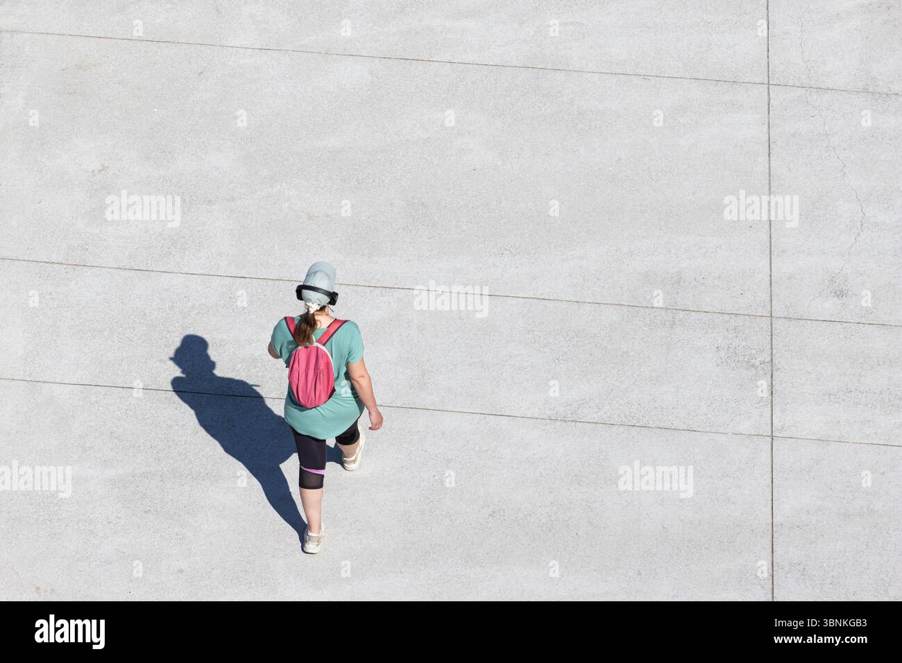 la donna curvy vestita di abbigliamento sportivo cammina con un cappello, cuffie e zaino su una superficie di cemento durante una giornata di sole. Spazio di copia vista ad angolo alto Foto Stock