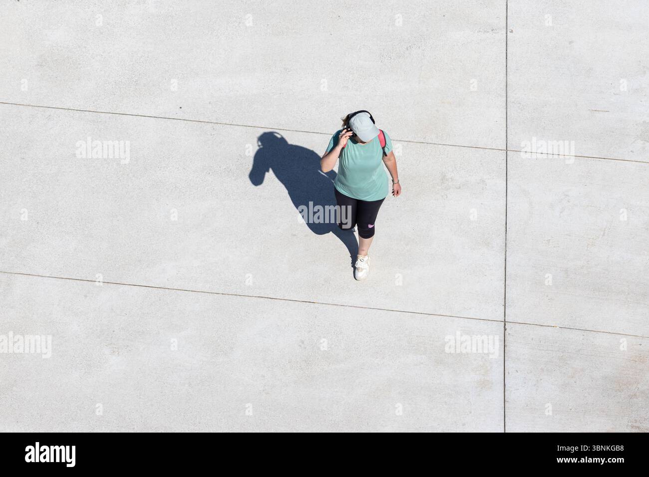 Donna che indossa un abbigliamento sportivo e un cappello con le cuffie mentre cammina su una passerella in cemento nelle giornate di sole. Spazio di copia, vista ad angolo alto Foto Stock