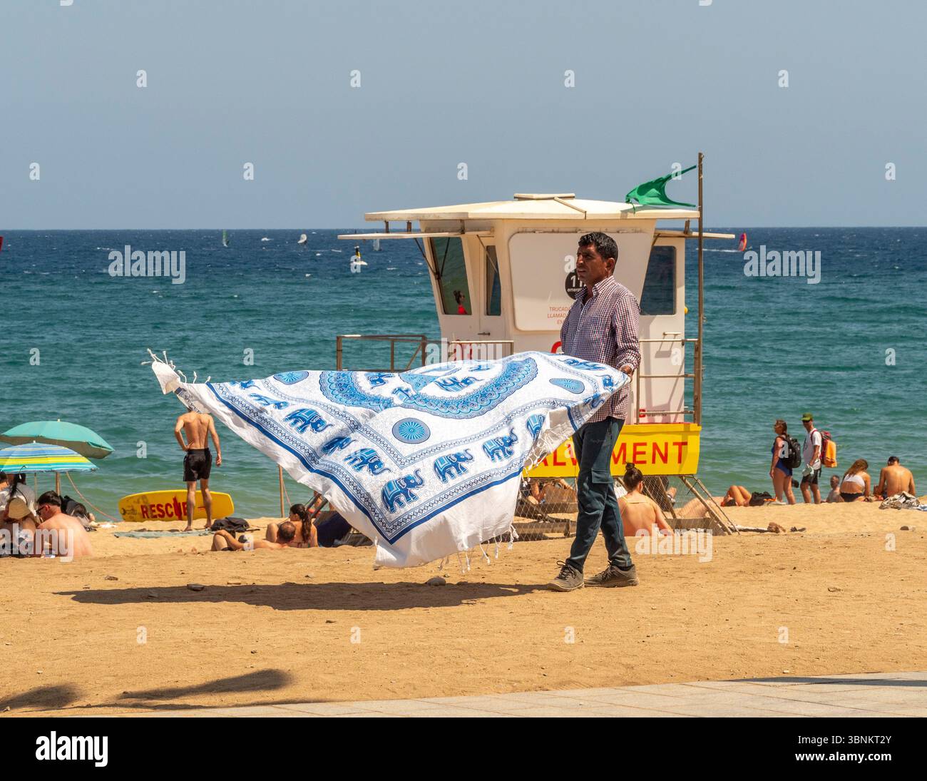 Un venditore di spiaggia cammina lungo la sabbia di una trafficata spiaggia di Barcellona, mostrando una grande coperta di elefante a motivi geometrici in vendita ai bagnanti. Foto Stock