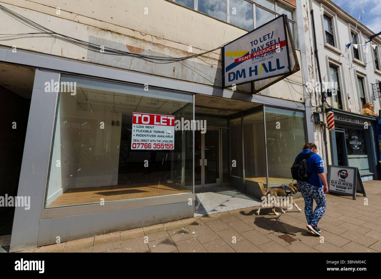 DUMFRIES, SCOZIA, Regno Unito - 19 giugno 2024 - Una donna e il suo cane passano davanti a un'unità di vendita vuota nella High Street di Dumfries, Scozia, Regno Unito. Come molti suc Foto Stock