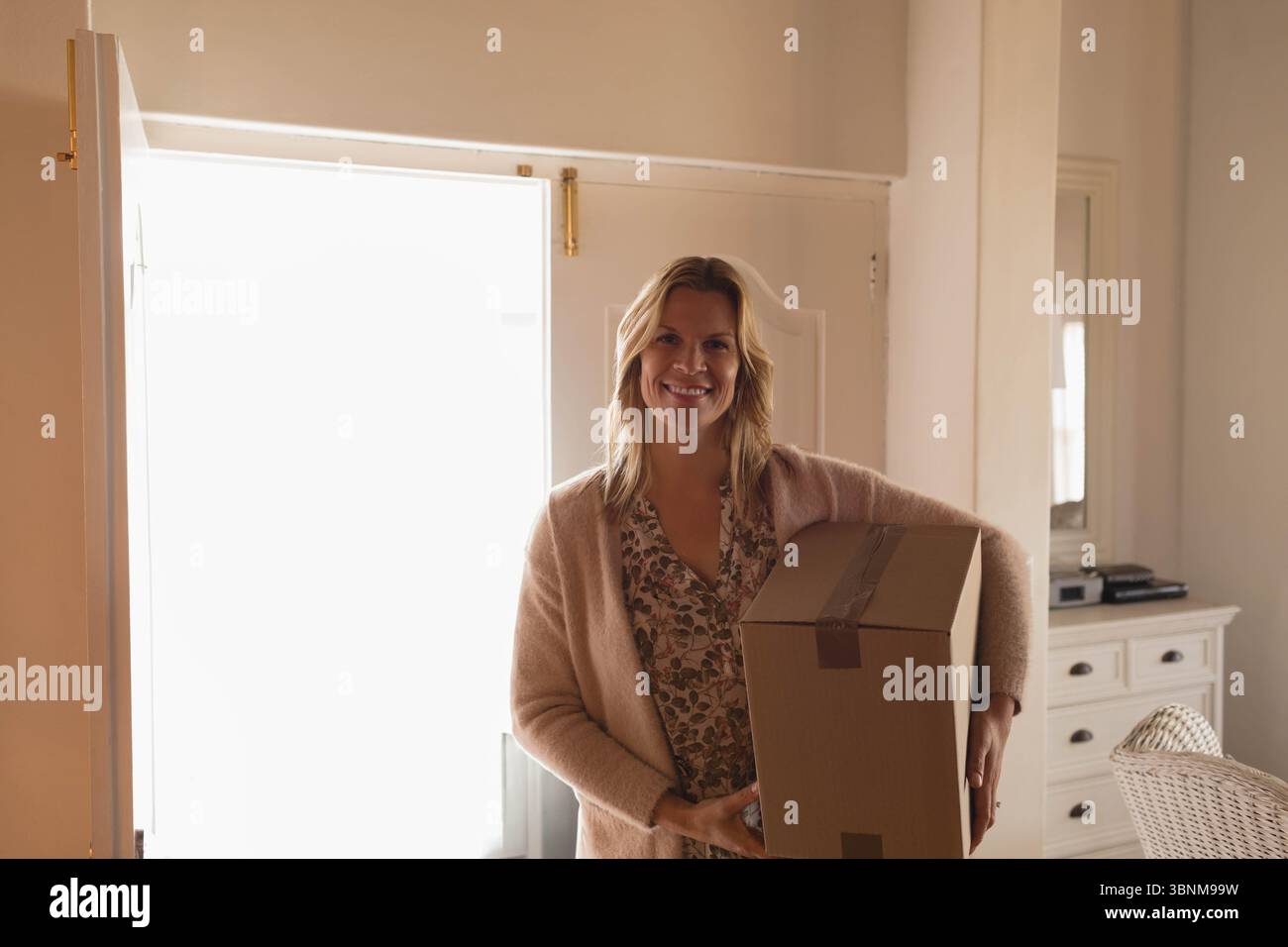 Donna in piedi vicino a una porta luminosa aperta all'ingresso, tenendo la scatola mobile accanto allo specchio del comò, sorridendo Foto Stock