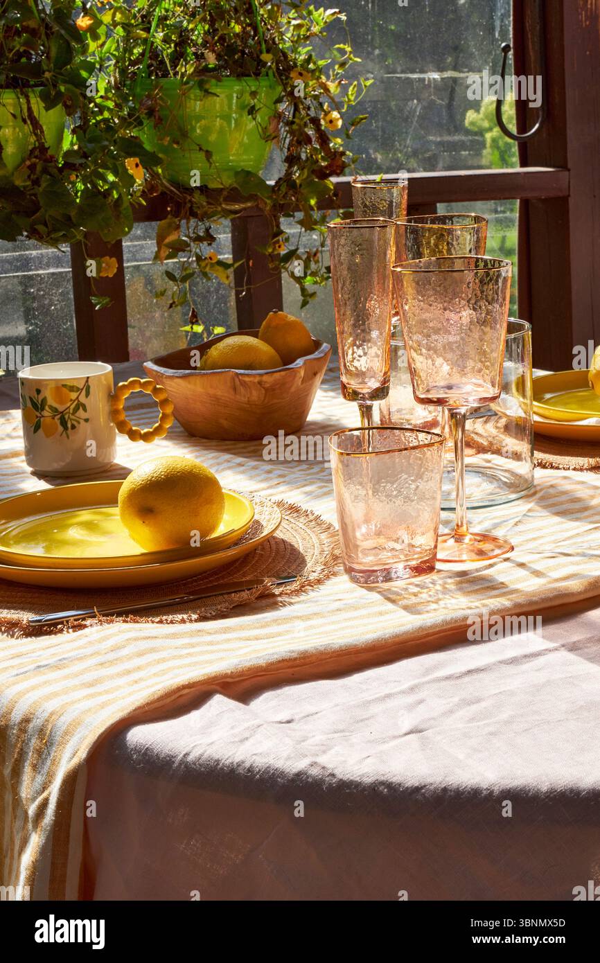 Un luminoso tavolo estivo decorato con limoni, ortensie verdi e bicchieri. La luce del sole si riversa attraverso le piante appese, creando un'atmosfera accogliente e allegra Foto Stock