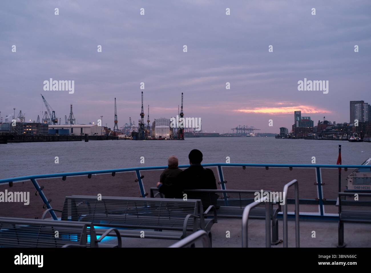 Padre e figlio sul ponte superiore di un traghetto HVV durante una traversata sull'Elba al crepuscolo con vista sul porto dei container Foto Stock