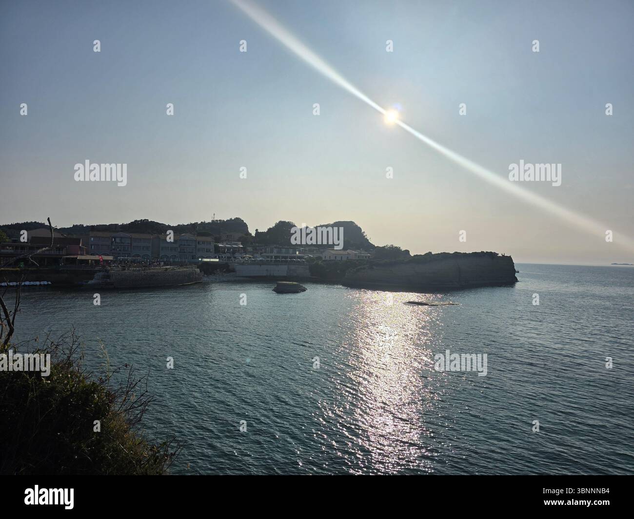 Mare calmo e blu che riflette la luce del sole, che conduce a una città costiera greca. Terreno collinare e cielo limpido. Foto Stock