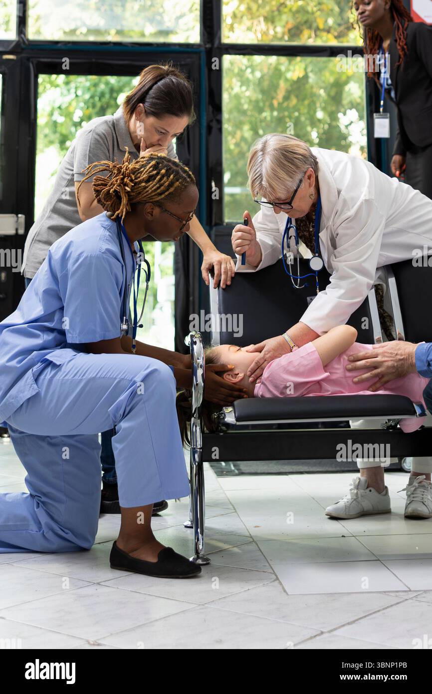 Madre scioccata in cerca di aiuto professionale per bambini incoscienti presso la hall della clinica, il team sanitario diversificato si mobilita per fornire cure critiche in una situazione di emergenza pediatrica salvavita. Foto Stock