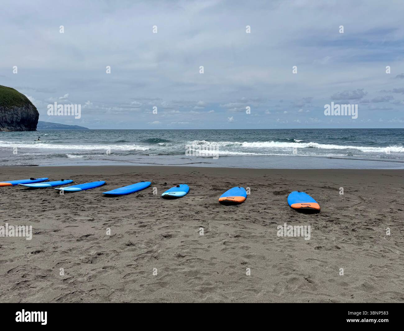 Una fila di tavole da surf sulla spiaggia di sabbia vulcanica dell'isola di São Miguel, Azzorre, Portogallo. Le onde dell'Oceano Atlantico si infrangono sotto un cielo parzialmente nuvoloso. - Immagine stock catturata con smartphone
