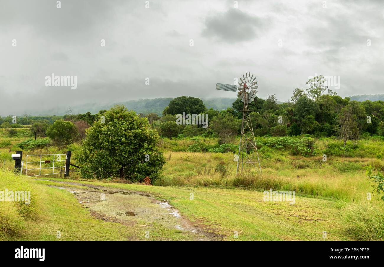 Scenario paesaggistico di una pompa dell'acqua ecologica con un cielo nuvoloso e una superficie bagnata dopo una tempesta di pioggia all'ingresso di una fattoria nell'Australia rurale. Foto Stock