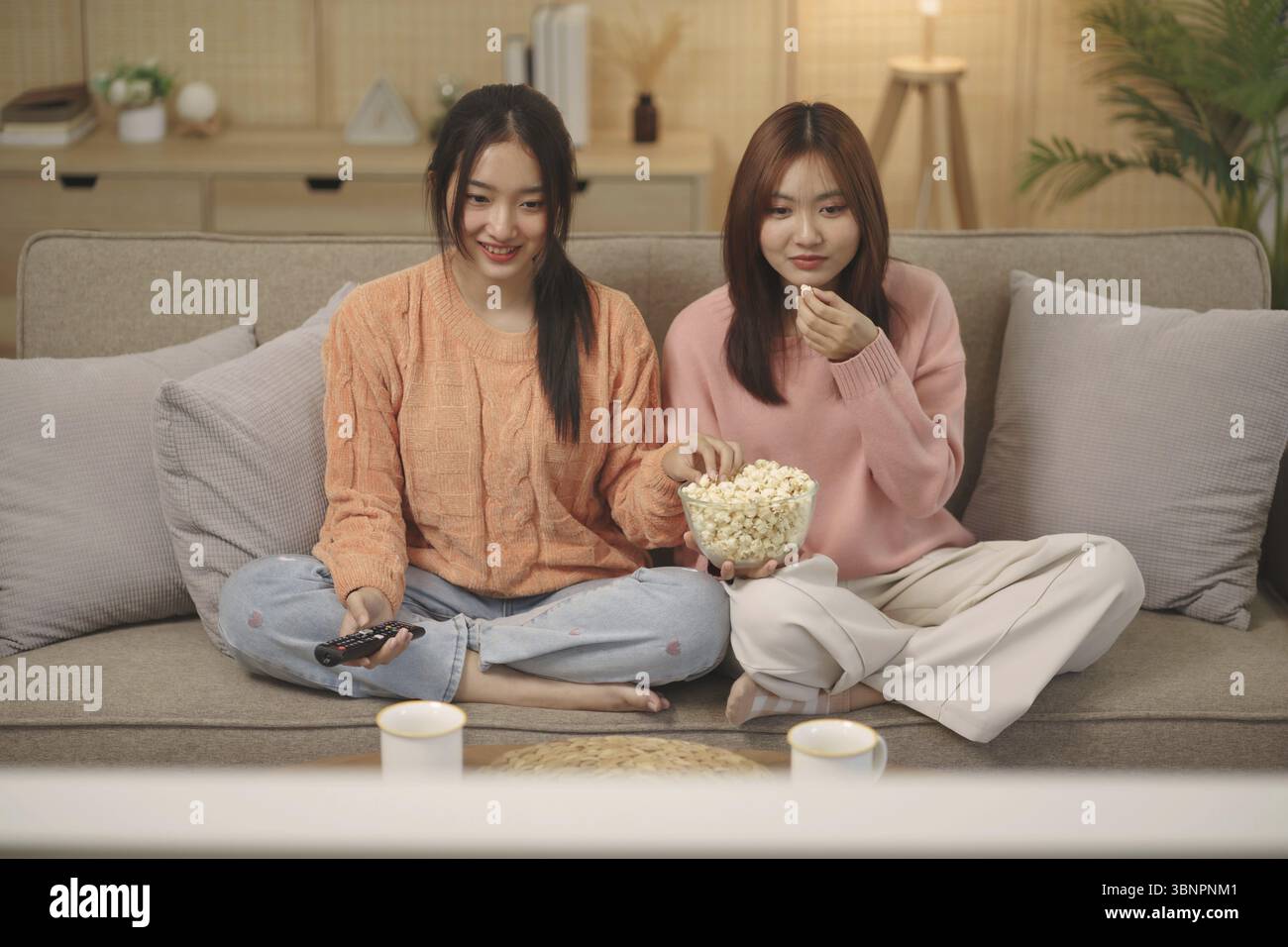 Amici felici ragazza asiatica che si diverte a guardare la televisione con popcorn usando il telecomando scegliendo il film della serie TV. Godersi la televisione rilassarsi a casa Foto Stock